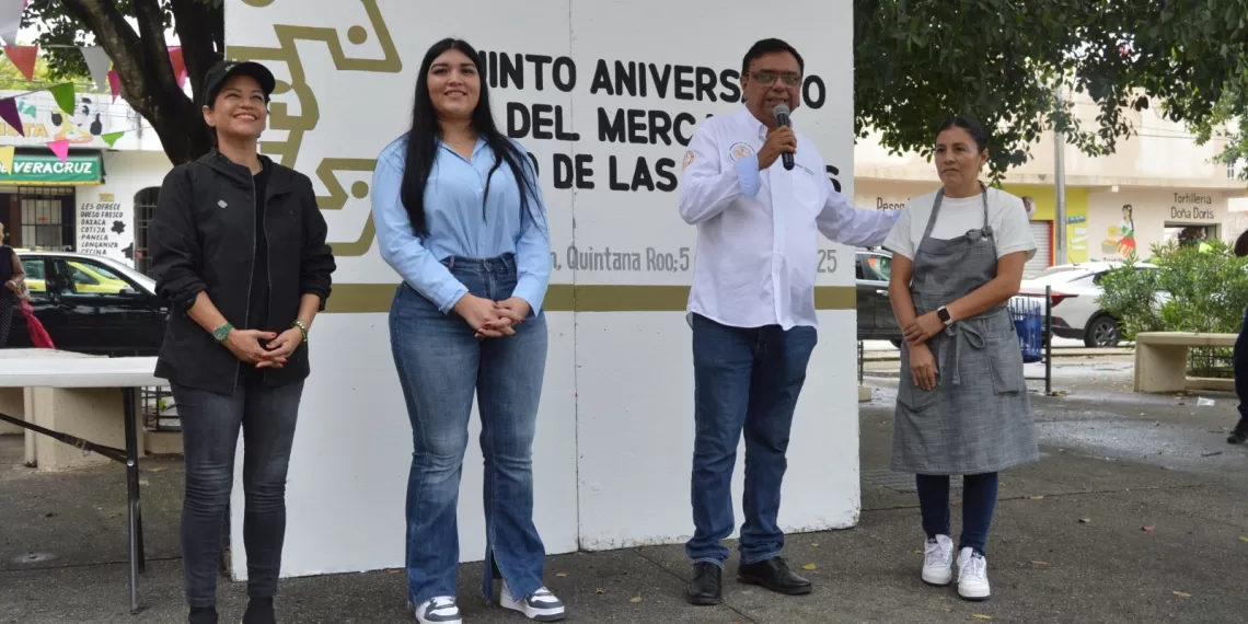 A group of four individuals, including a man speaking into a microphone, stands in front of a sign celebrating the fifth anniversary of the Mercado de las Flores. Two women are smiling, and one is dressed in a gray apron. The setting appears festive with colorful decorations in the background.$#$ CAPTION