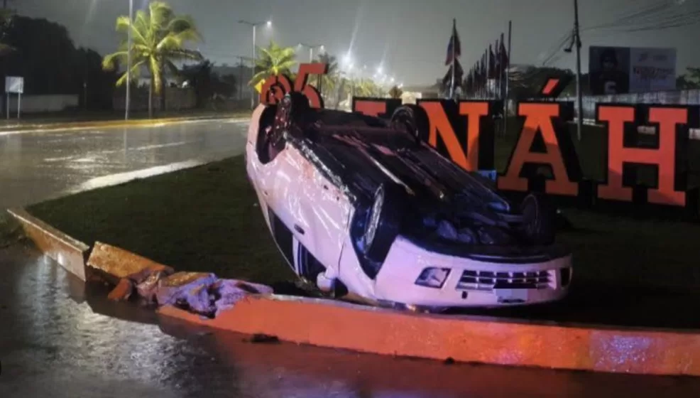 A white car overturned in a flooded area during a rainstorm, with large letters in the background indicating a location name