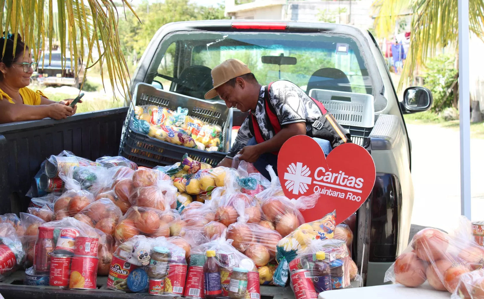 A man arranges bags of food in the back of a pickup truck while a woman uses her phone nearby. The truck features a heart-shaped sign for Caritas Quintana Roo. $#$ CAPTION