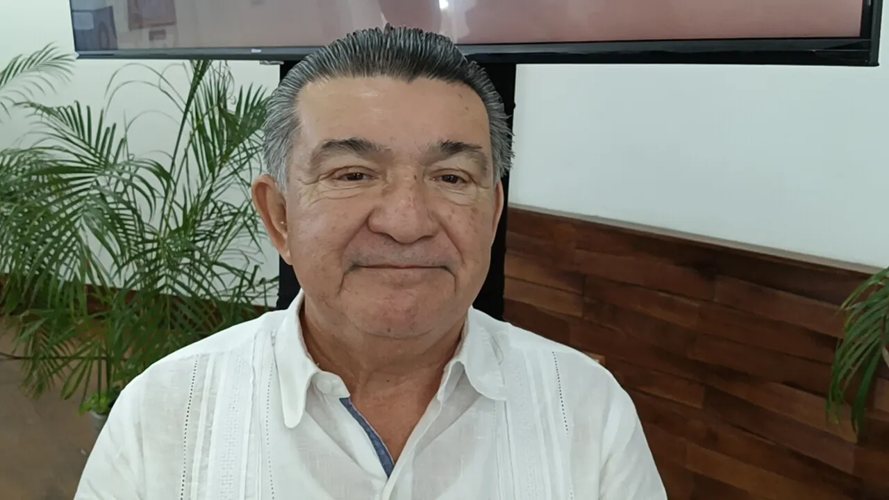 An older man with a slight smile, wearing a white shirt, sitting in front of a wooden wall with some greenery in the background.