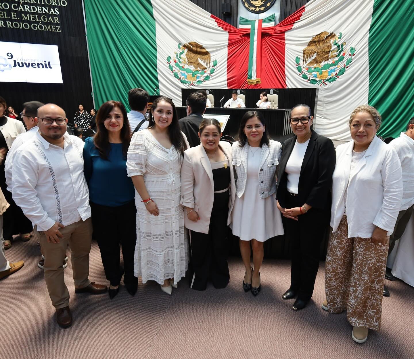 A group of people posing for a photo in front of the Mexican flag in a legislative chamber setting.$#$ CAPTION