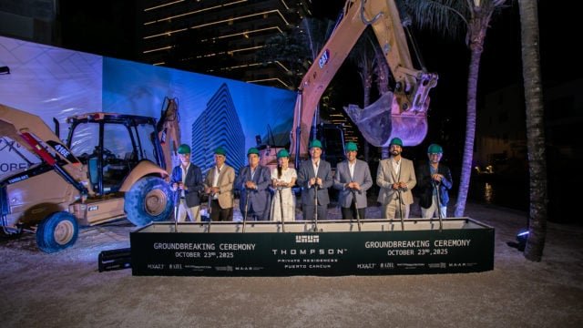 Seven individuals in formal attire and hard hats pose for a groundbreaking ceremony in front of construction machinery and a banner.$# CAPTION