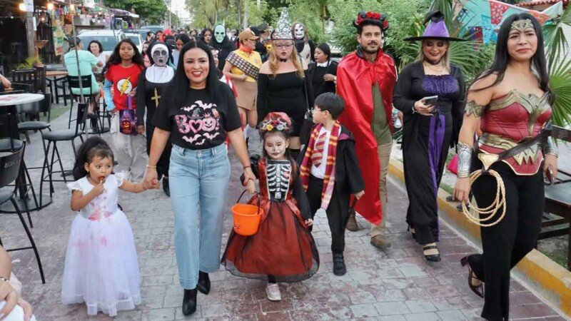 A lively Halloween parade featuring families and children in a variety of costumes, walking down a vibrant street decorated for the occasion.$#$ CAPTION