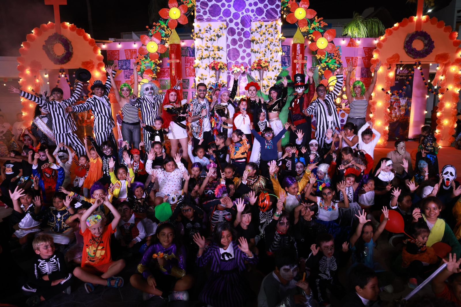 A large group of children and performers in Halloween costumes, gathered together with festive decorations in the background. Many kids are cheering and raising their hands.