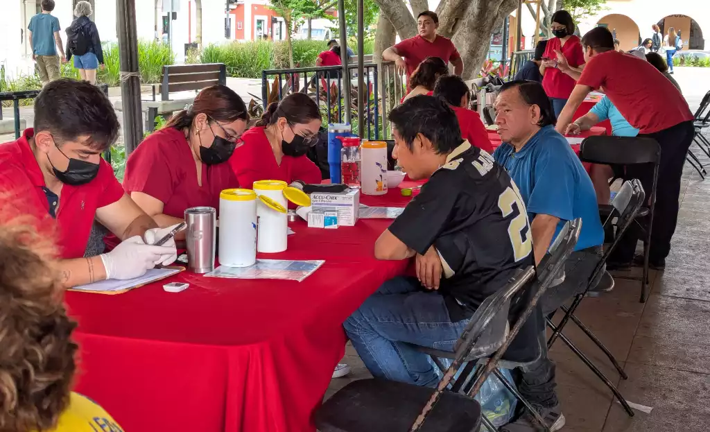 health workers in red shirts and masks seated at a table engaged with community members in a public space