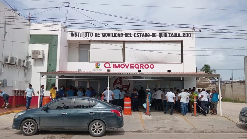 A busy scene outside the Instituto de Movilidad del Estado de Quintana Roo, showing a crowd of people gathered in front of the building with a parked car in the foreground.$#$ CAPTION