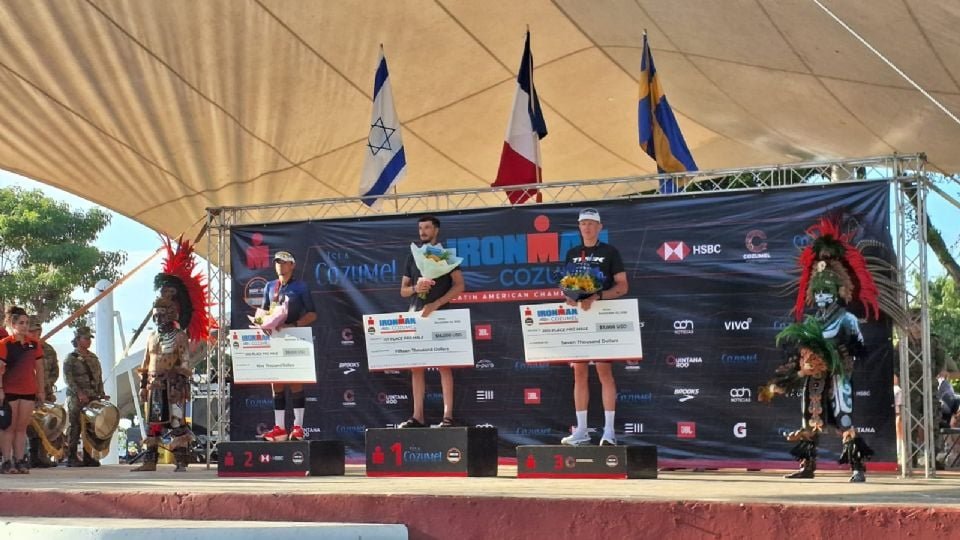 Athletes on stage at the Ironman Cozumel awards ceremony holding trophies and checks with a cultural performer in traditional attire nearby