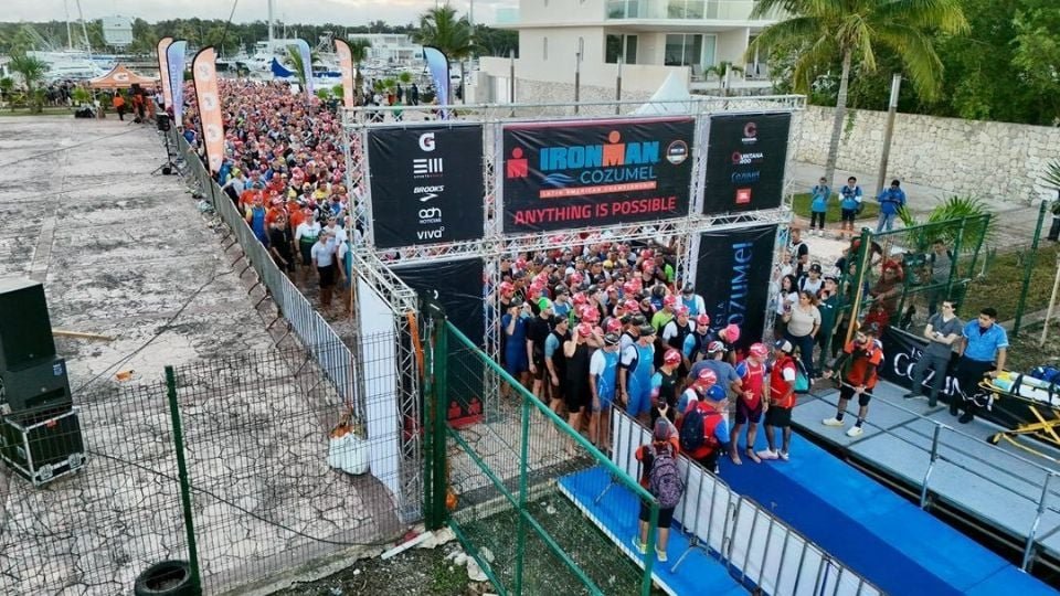 A large group of triathletes gathered at the start line of the Ironman Cozumel event wearing colorful swim caps and wetsuits