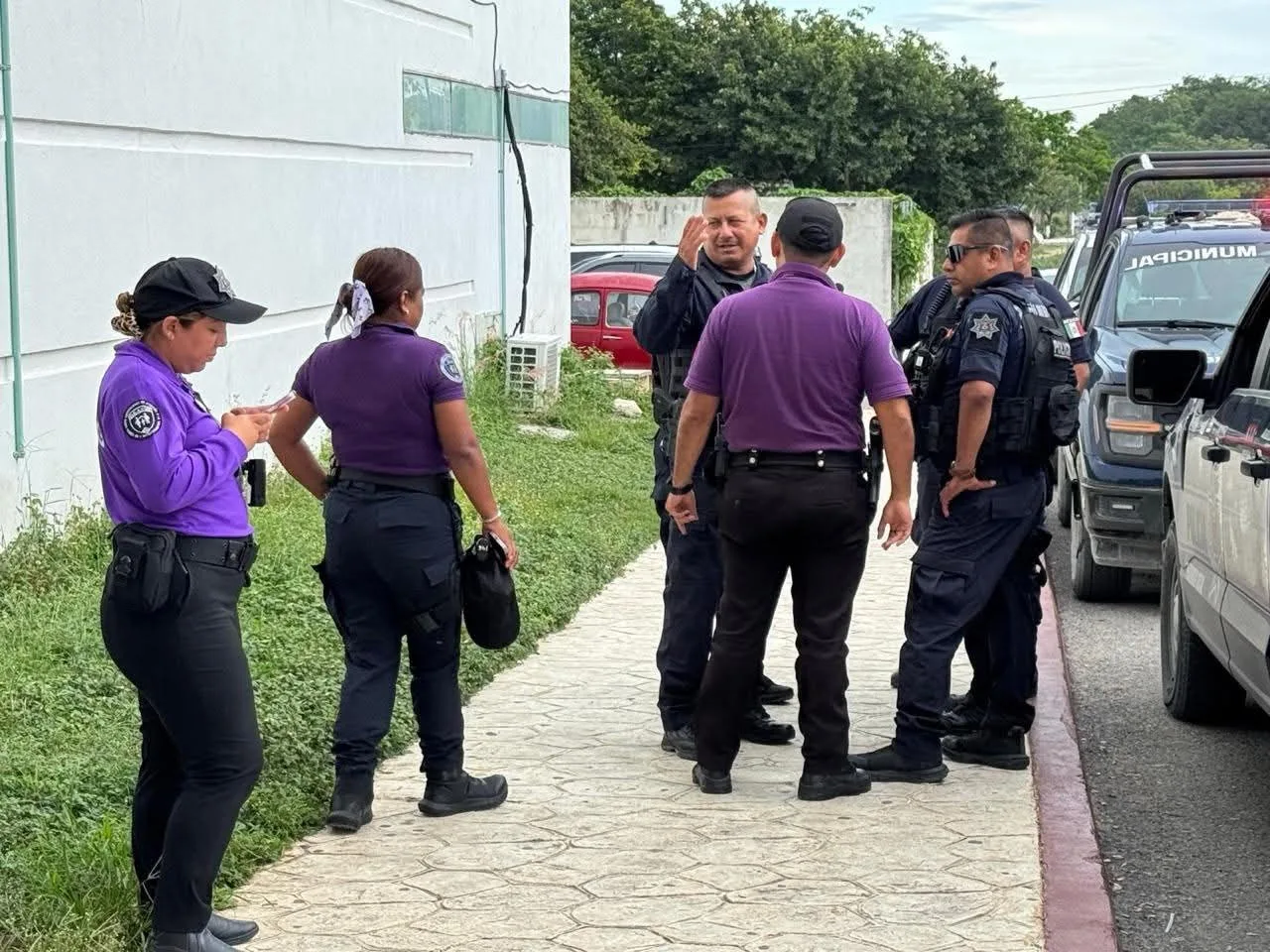 A group of police officers in various uniforms, including purple shirts, engage in conversation on a street while one officer checks their phone. A police vehicle is visible in the background.