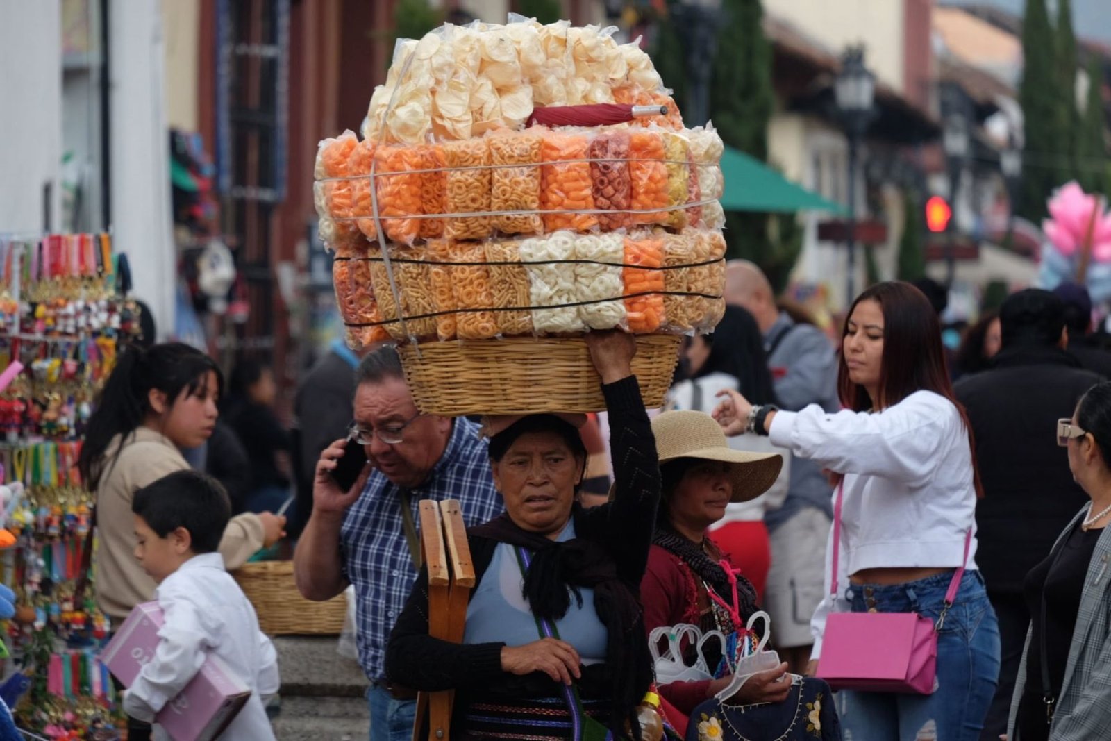 A woman carrying a large basket of colorful snacks on her head while walking through a crowded market. Several other people are visible nearby, engaged in shopping or conversation.