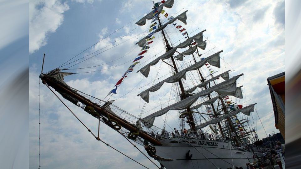 a tall ship adorned with flags and sails against a cloudy sky with people on board and nearby-01112025