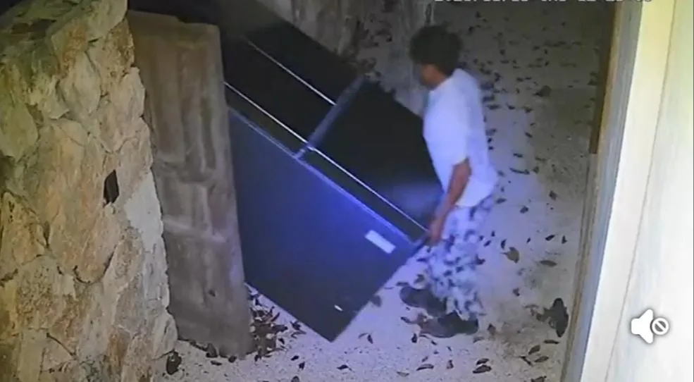 A person is lifting a large dark object in a dimly lit area with scattered leaves on the ground.