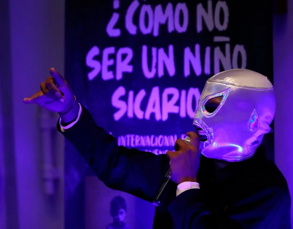 a performer wearing a silver mask holding a microphone while gesturing with one hand in front of a dark background with text-$# CAPTION