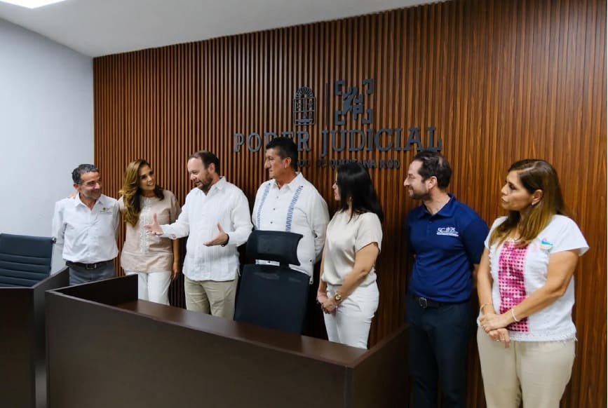 A group of six individuals standing together in a modern office setting, with a wooden wall featuring the words 'PODER JUDICIAL' in the background. They appear to be engaged in conversation and collaboration.$#$ CAPTION