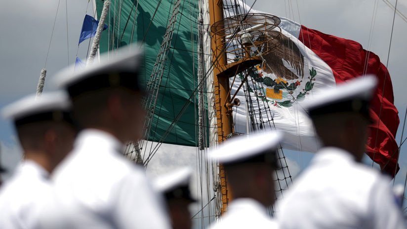 Mexican naval officers in uniforms stand in formation with the Mexican flag and a green flag in the background.