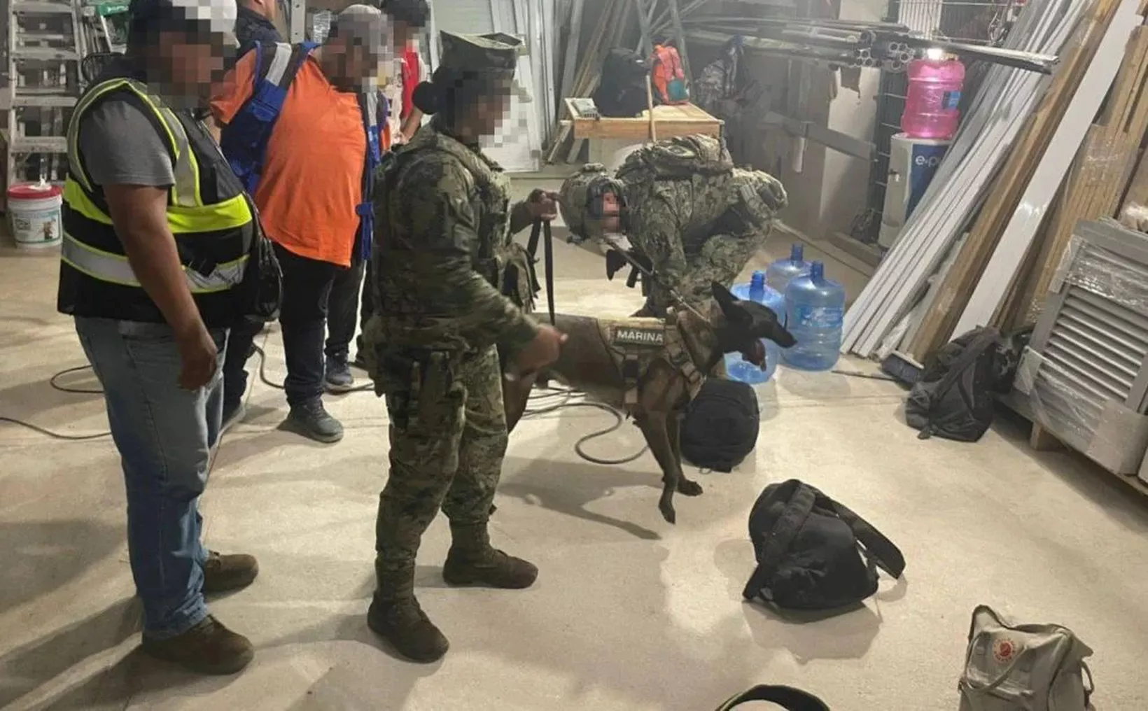 Military personnel and a service dog in a warehouse setting, preparing for a mission or operation.$# CAPTION