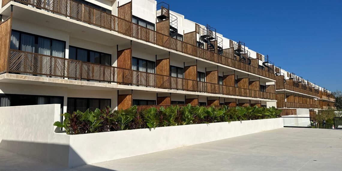 A modern residential building with balconies and greenery in front under a clear blue sky