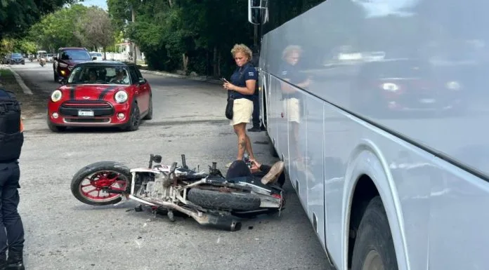 A motorcycle lies on its side near a bus, with a woman standing nearby looking at her phone, and a red car in the background.$#$ CAPTION
