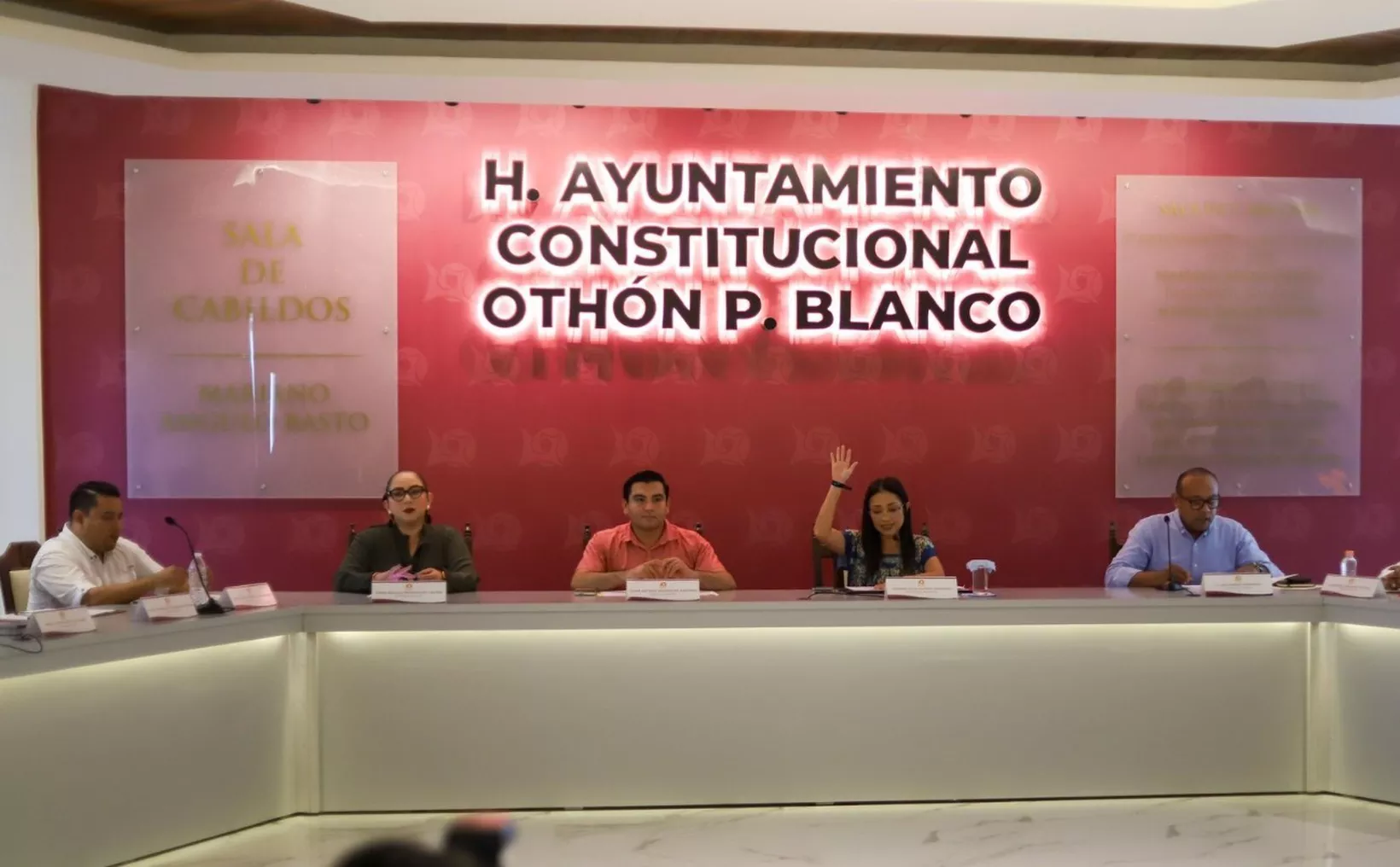 A panel of officials seated at a table during a meeting at the municipal council in Othón P. Blanco, with one female participant raising her hand to speak.$# CAPTION