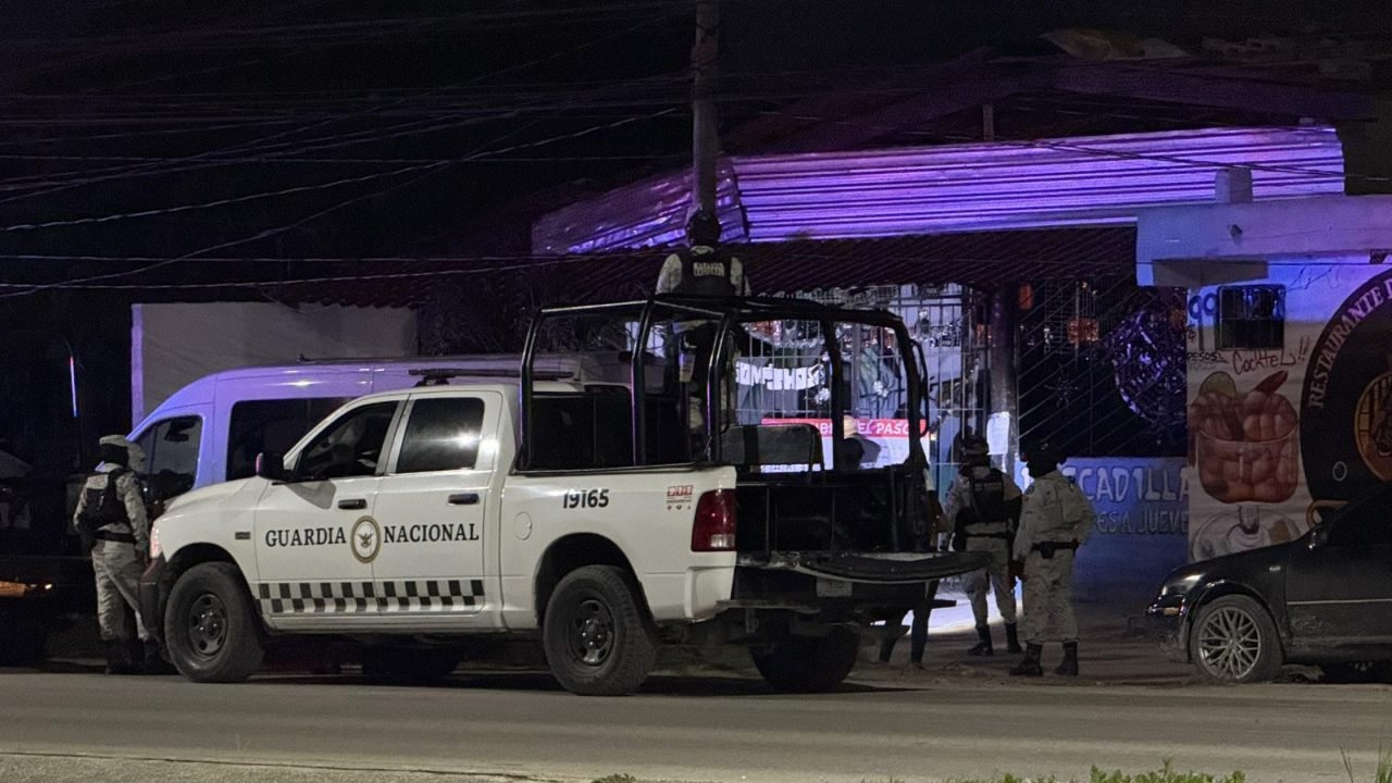 A National Guard truck is parked on a street at night, with personnel engaging in an operation near a building illuminated by purple lights.$#$ CAPTION