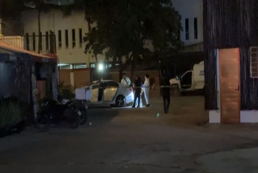 A nighttime scene showing a group of people near a car, with some officers inspecting it in an urban area. There is a police vehicle in the background and a building nearby. Light reflects off surfaces creating a dramatic atmosphere.$#$ CAPTION
