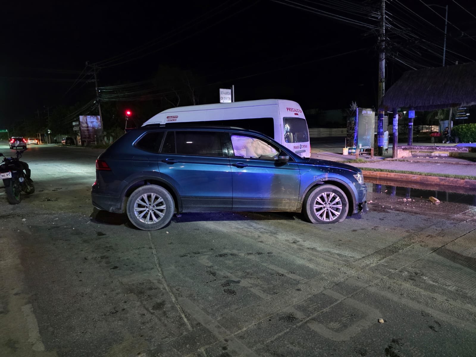 A dark blue SUV is parked at the side of a road at night, with a van visible nearby and traffic signals in the background.