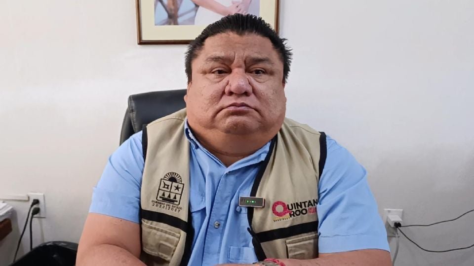a man wearing a beige vest with a logo seated at a desk in an office -$# CAPTION