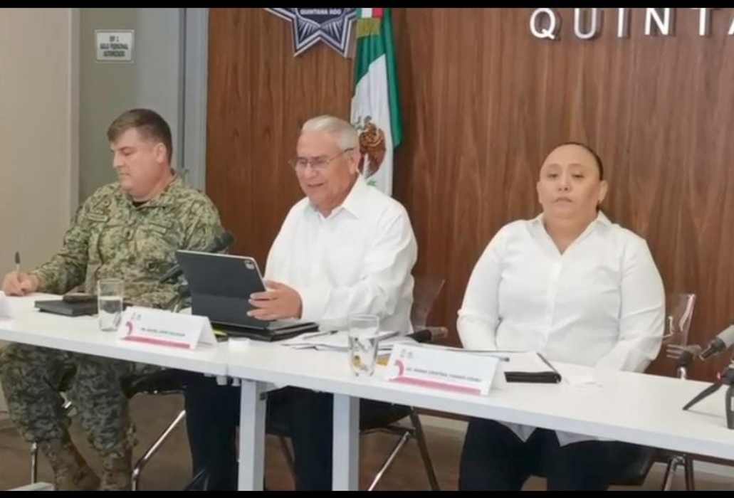 A meeting of officials in a conference room, with a military officer, a man in glasses, and a woman seated at a table. Mexican flags are displayed in the background.$#$ CAPTION