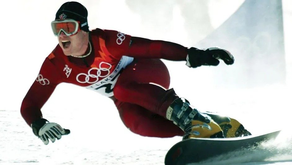 A snowboarder wearing a red Olympic uniform competes on a snowy slope, leaning into a turn with intense focus and determination.