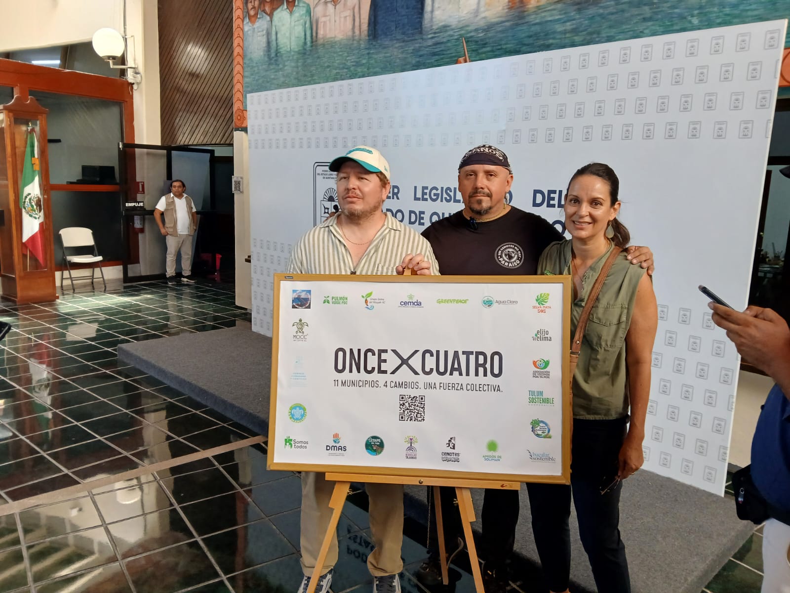 Group of individuals holding a sign for the Once x Cuatro initiative, promoting collaborative changes in 11 municipalities. The background features a Mexican flag and a mural.-$# CAPTION
