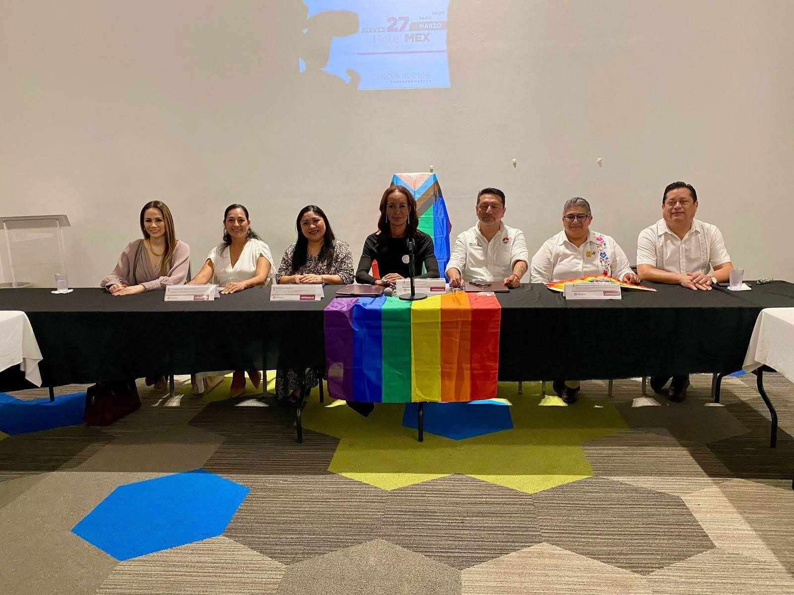 A panel of seven speakers seated behind a table with a rainbow flag, discussing LGBTQ+ rights.$# CAPTION