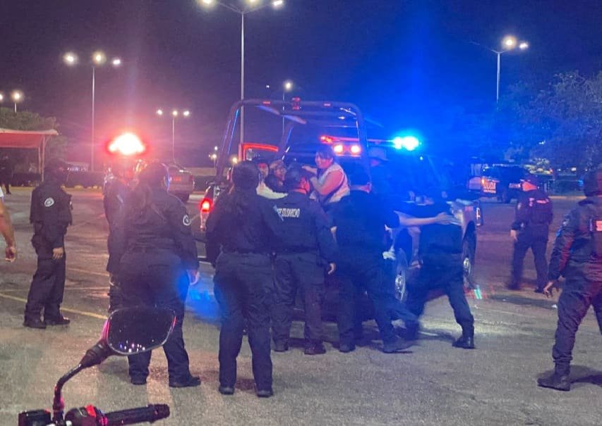 A group of police officers is responding to an incident at night, with flashing lights in the background. Several individuals are engaged in an activity near a police vehicle.