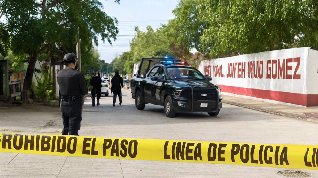 A police officer stands near a police line at a crime scene, with a patrol vehicle and officers in the background. Urban setting with greenery and wall graffiti present.