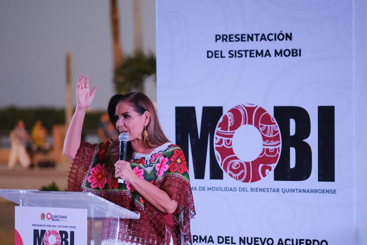 A speaker in traditional clothing gestures while presenting the MOBI system at an event in Quintana Roo, with a large banner in the background.$# CAPTION