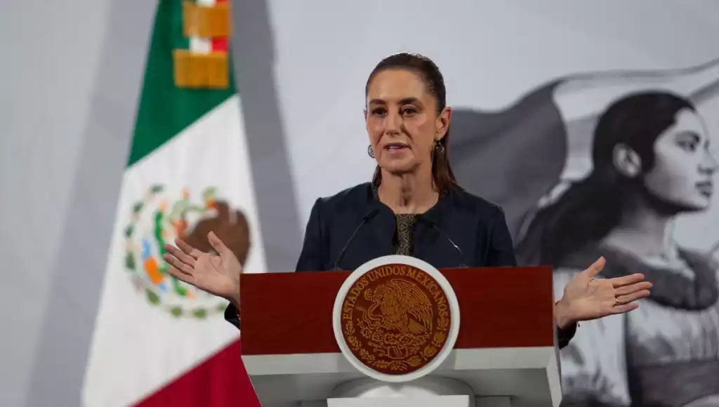 a woman speaking at a podium during a press conference with the Mexican flag in the background