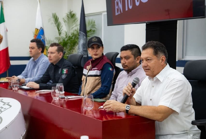 A group of five speakers at a press conference discussing community initiatives, with flags in the background and a large screen displaying a message.$#$ CAPTION