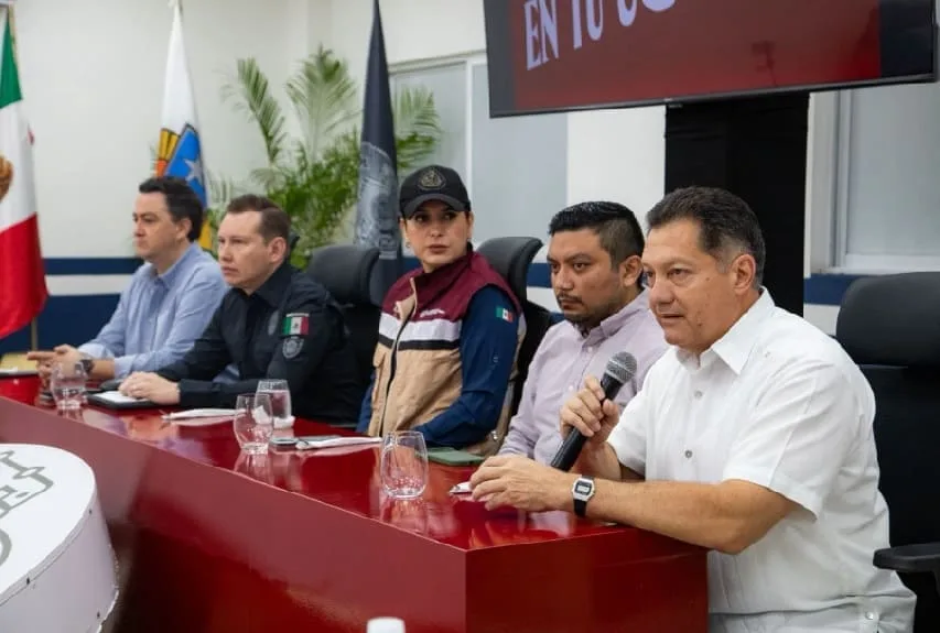 A group of five speakers at a press conference discussing community initiatives, with flags in the background and a large screen displaying a message.$#$ CAPTION