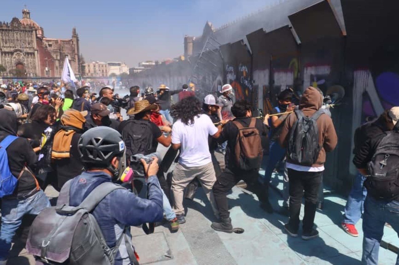 A large crowd of protesters engaged in a tense confrontation with authorities in Mexico City, with smoke and chaos in the background.$# CAPTION