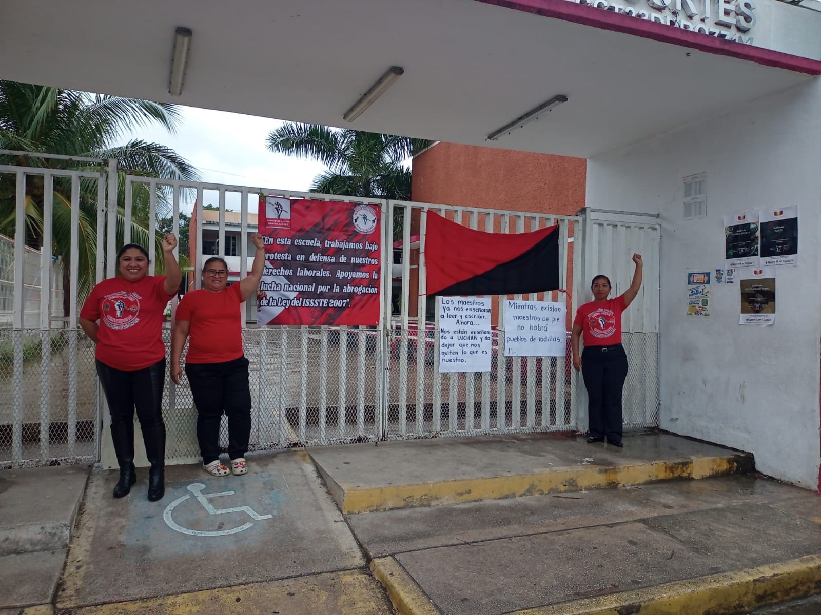 Two women and one woman raising fists in solidarity in front of a school gate with banners and a flag