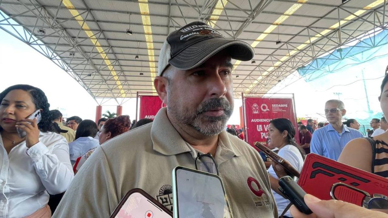 A man in a baseball cap speaks to the media at a public event, surrounded by individuals using smartphones and engaging in conversation under a large structure.$# CAPTION