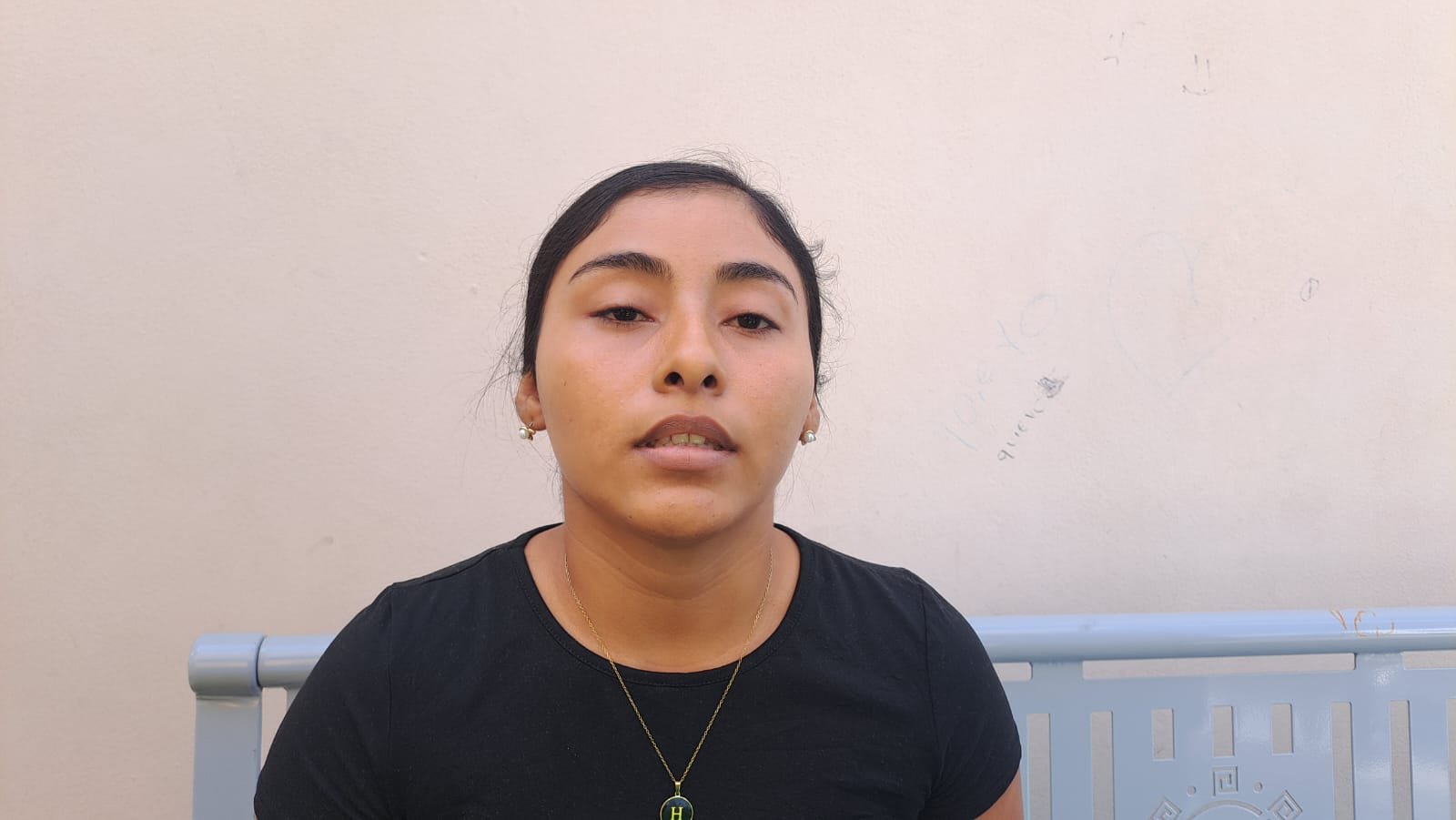 A young woman with black hair wearing a black t-shirt and a necklace, sitting on a bench against a plain wall. She appears thoughtful and serious.