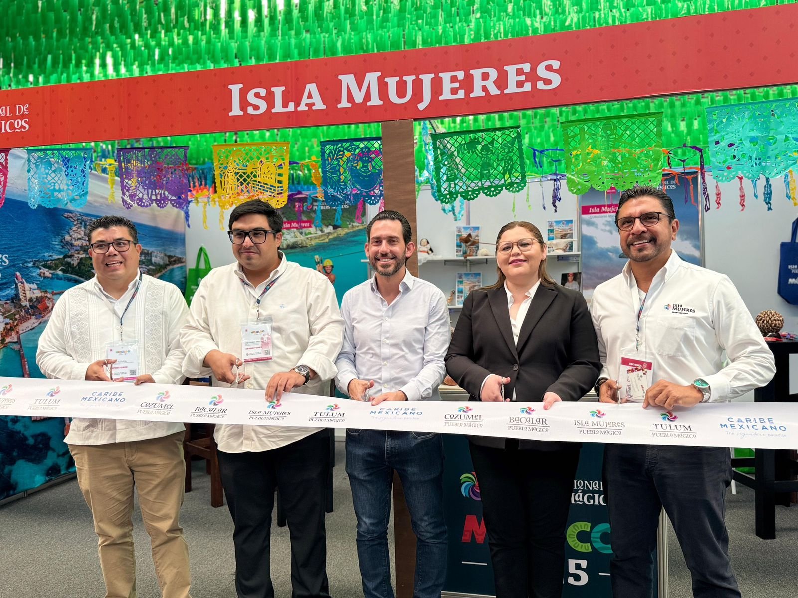 A group of five individuals participating in a ribbon-cutting ceremony at the Isla Mujeres tourism booth, showcasing a backdrop of vibrant decorations and promotional materials.$# CAPTION