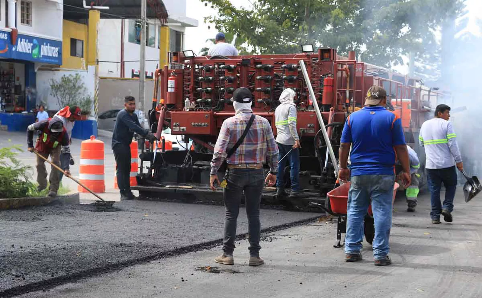 Workers laying asphalt on a road with machinery and tools in use, surrounded by construction cones and a busy street