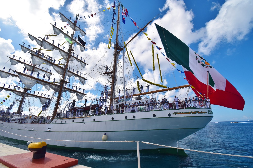 A majestic tall ship adorned with flags and the Mexican flag, sailing in clear blue waters under a partly cloudy sky.$# CAPTION