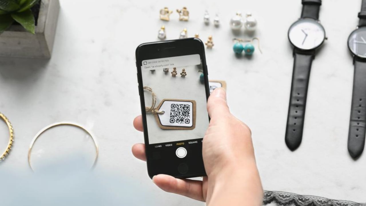 A person using a smartphone to scan a QR code on a tag, with jewelry items arranged on a table in the background.$# CAPTION