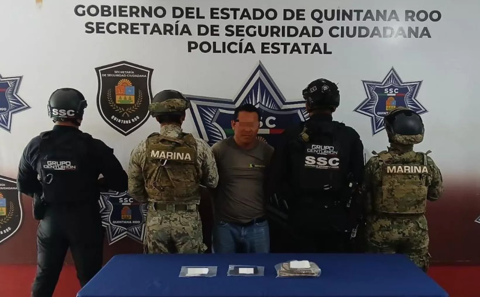 A suspect is surrounded by law enforcement officers during a security operation in Quintana Roo, Mexico, with a table displaying evidence in front of them.$# CAPTION