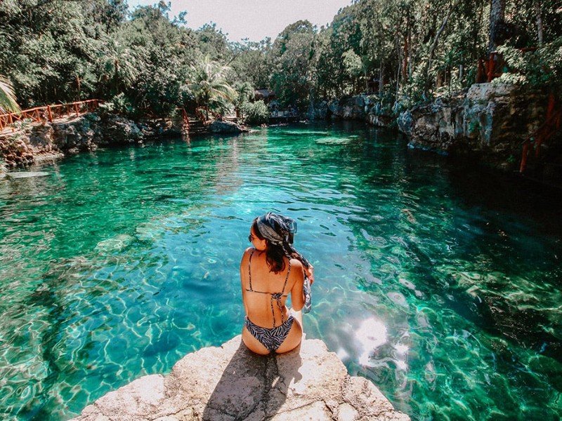 A person sitting on a rock by a turquoise body of water surrounded by lush greenery