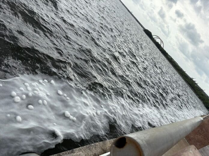 A calm body of water with white foam patterns along the surface, viewed from an elevated angle. A pipe is visible in the foreground. The sky is partly cloudy.$#$ CAPTION