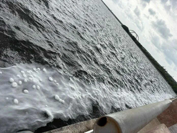 A calm body of water with white foam patterns along the surface, viewed from an elevated angle. A pipe is visible in the foreground. The sky is partly cloudy.$#$ CAPTION