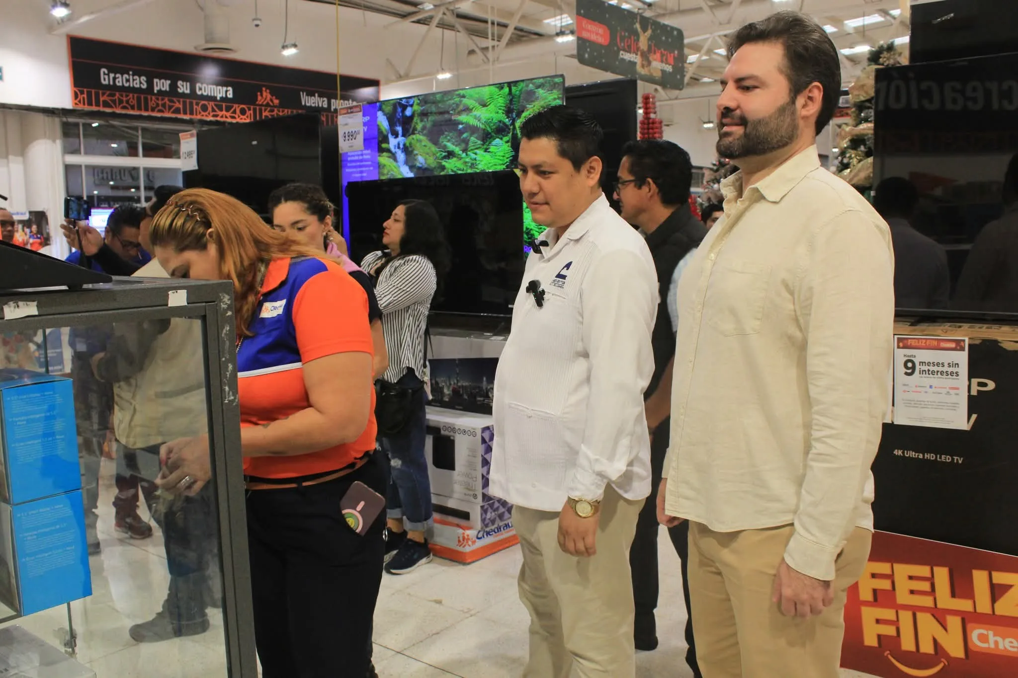 A woman in an orange and blue shirt is interacting with a display case in an electronics store, while two men and several other shoppers look on in the background. The store features televisions and promotional signage.